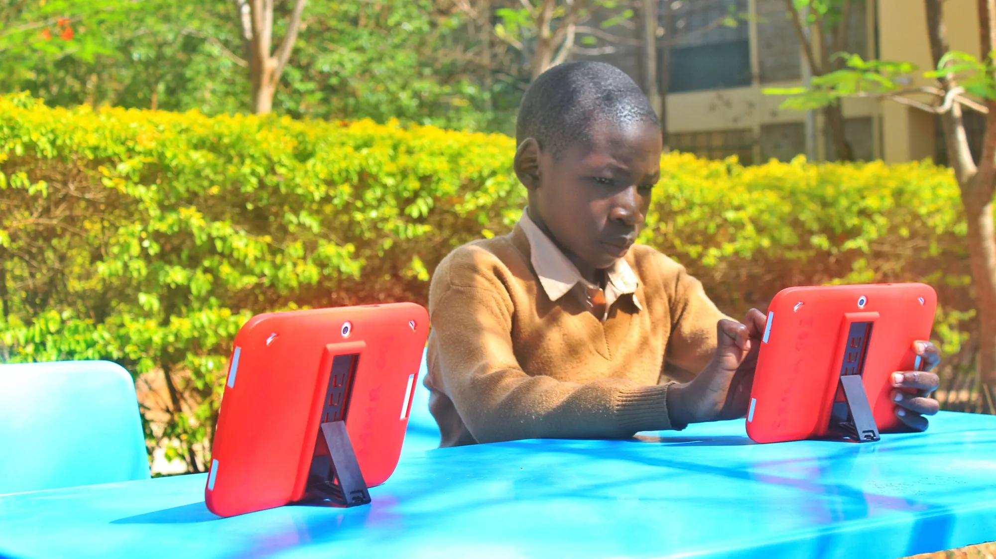 A girl in a beige uniform using a red tablet at a blue table outdoors